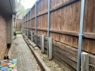 Deteriorating railroad tie retaining wall with steel post supports along a brick paver path, awaiting replacement with Milsap stone in DFW
