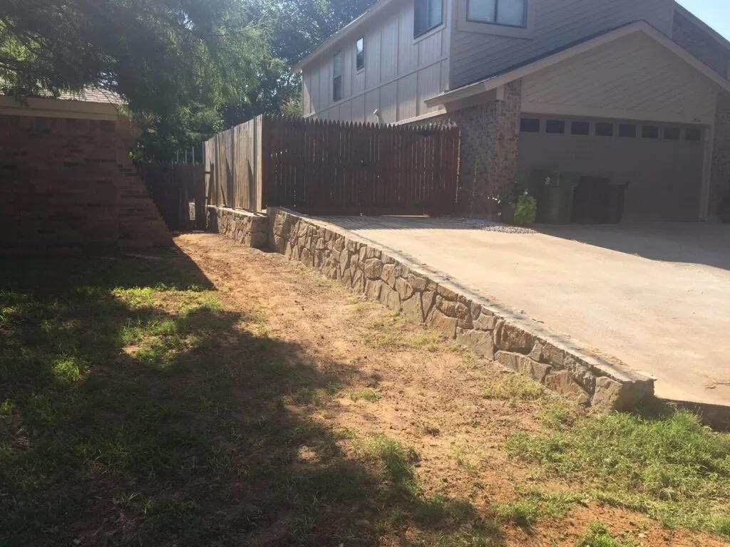 Natural stone retaining wall running alongside a residential driveway in Grapevine TX, holding back a sloped yard next to a two-story brick home