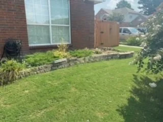 Low stone retaining wall along brick home's front yard in Dallas, holding a garden bed with green lawn and wood fence in background