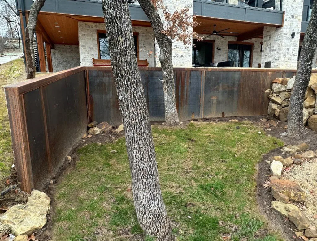 Cor-Ten steel panel retaining wall with rusted patina supporting a sloped backyard below a modern stone home in Colleyville TX