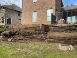Failing Railroad Tie Retaining Wall Replacement | Colleyville TX Deteriorating railroad tie retaining wall with rotting timbers leaning and collapsing behind brick home in DFW before replacement