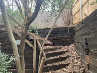 Deteriorating railroad cross-tie retaining walls and wooden staircase surrounded by trees in a Colleyville TX backyard, awaiting replacement with Milsap stone.