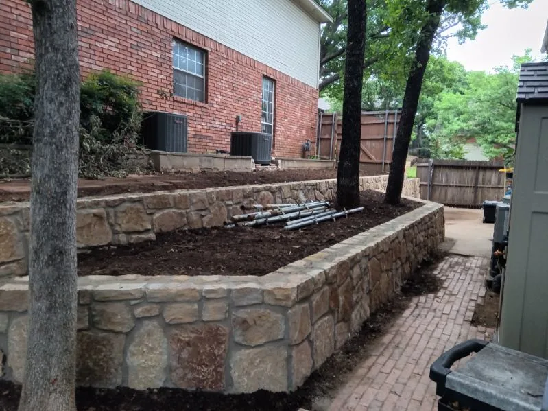 Tiered Milsap stone retaining walls replacing old cross tie wall in Grapevine TX backyard, with freshly graded soil beds alongside brick paver path