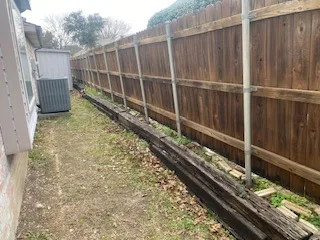 Deteriorating railroad tie retaining wall along a wood privacy fence in a DFW backyard, showing rotting timbers and soil erosion before Milsap stone replacement