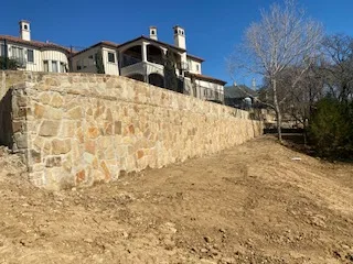 Large natural stone retaining wall supporting a luxury home's hillside lot, freshly graded soil at base, DFW area