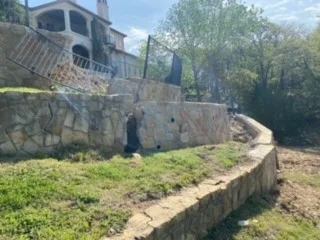 Worker demolishing deteriorating natural stone retaining wall beside upscale home, cleared dirt grade visible, preparing for new poured concrete wall in DFW.