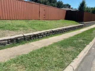 Concrete Block Retaining Wall Replacement – Carrollton TX Low concrete block retaining wall along a sloped residential yard beside a sidewalk and wood fence in Carrollton TX