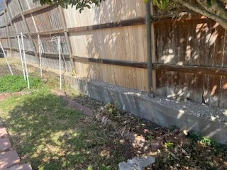 Deteriorating concrete retaining wall with cracked surface and exposed aggregate along wood privacy fence in Dallas backyard before replacement