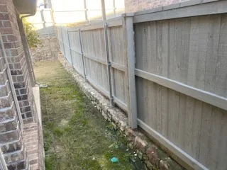 Side Yard Before Stone Wall Extension – Colleyville TX Narrow side yard between brick home and wood privacy fence showing low deteriorating stone base wall before Milsap stone extension and leveling work in DFW
