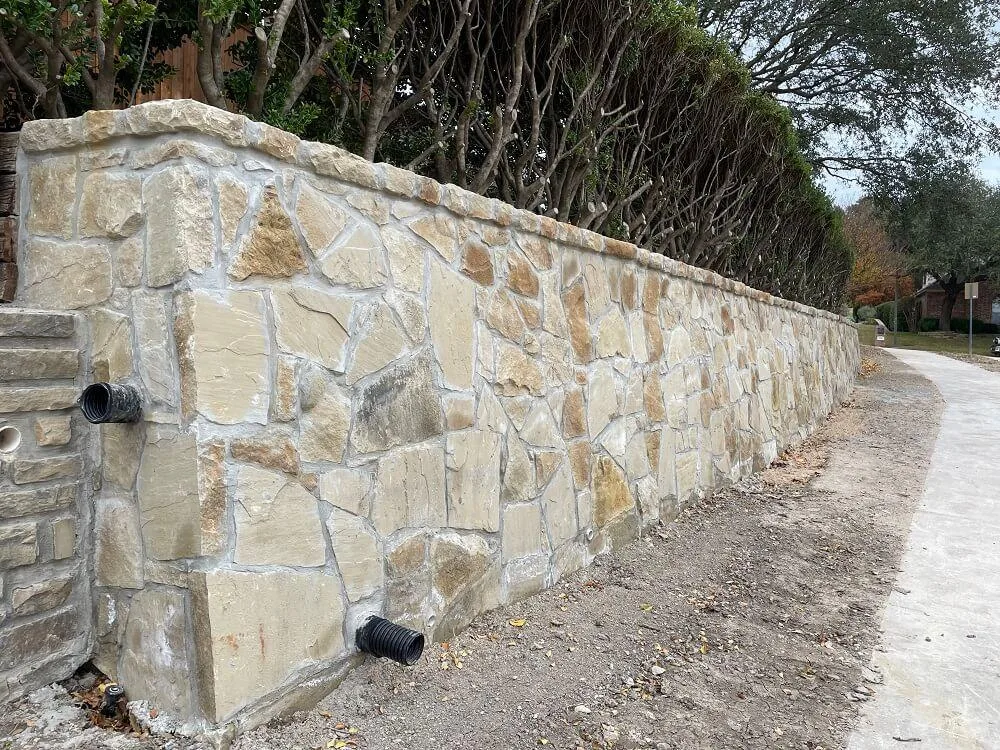 Long Granbury stone retaining wall with drainage pipes along a residential driveway, built to manage slope and erosion in DFW clay soil.