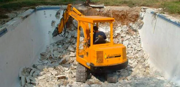 Yellow mini excavator breaking up concrete pool shell during residential pool removal demolition in DFW