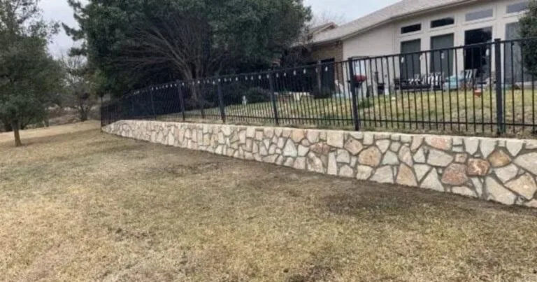 Completed Milsap stone retaining wall replacing a failed crosstie wall along a residential backyard grade in Carrollton, TX, with black iron fence above.