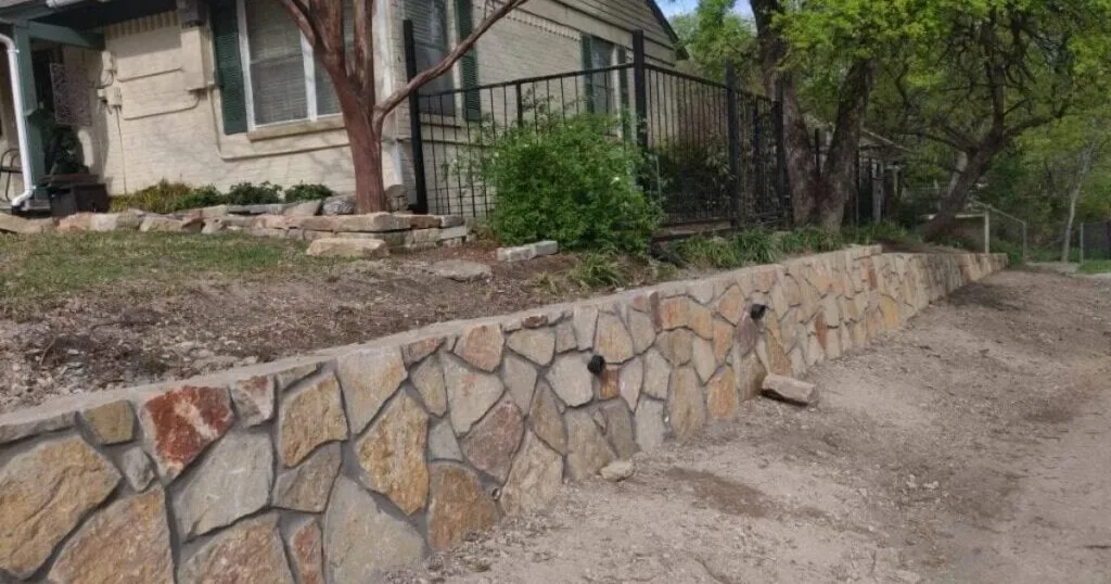 Newly built Granbury stone retaining wall along a residential front yard in Dallas, TX, with drainage weep holes and black iron fence above