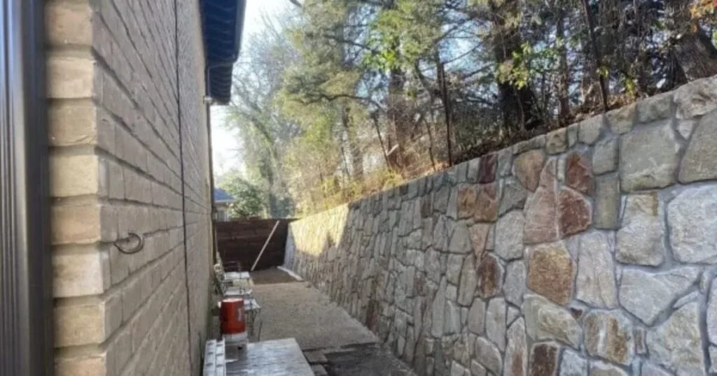 Newly built Granbury stone retaining wall running alongside a Dallas home, replacing a deteriorated wood wall, with trees visible above