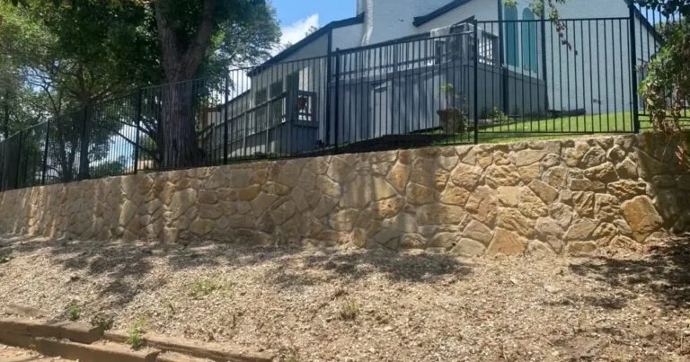 Completed Milsap stone retaining wall replacing old cross-tie wall on sloped residential yard in Irving, TX, with black iron fence above