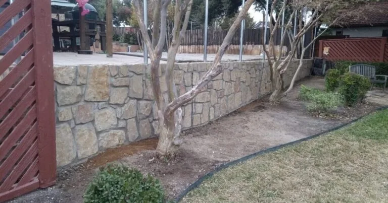 Newly built Milsap stone retaining wall separating a backyard patio from a lower lawn area in Irving, TX, with crape myrtle trees growing alongside.
