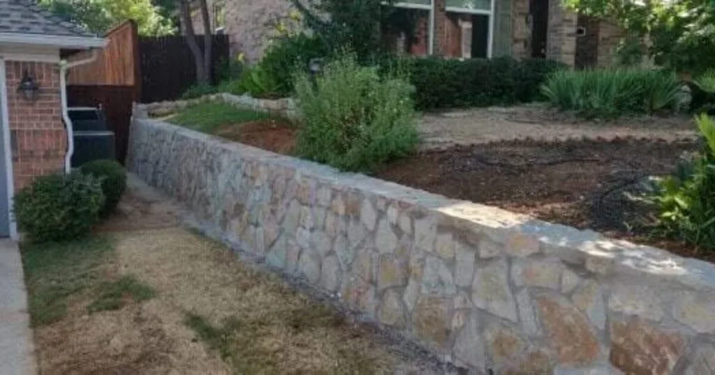 Newly built natural stone retaining wall along a residential driveway in Dallas–Fort Worth, holding raised garden bed with fresh mulch and plantings