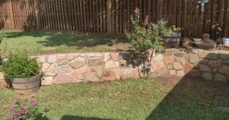 Natural stone retaining wall with warm tan and rust tones curving along a backyard in Carrollton TX, with planter barrels and shrubs above