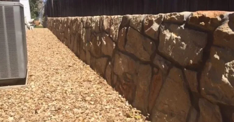 Natural stone retaining wall with large fitted Granbury-style blocks along a gravel yard beside a wood fence in DFW