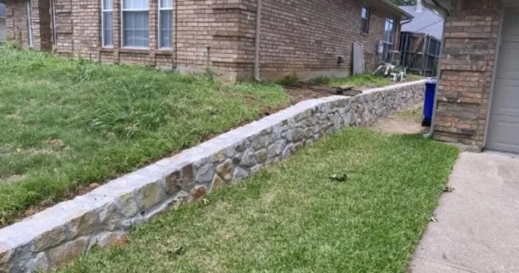 New natural stone retaining wall along a sloped front yard beside a brick home in Euless TX, controlling erosion near the driveway.