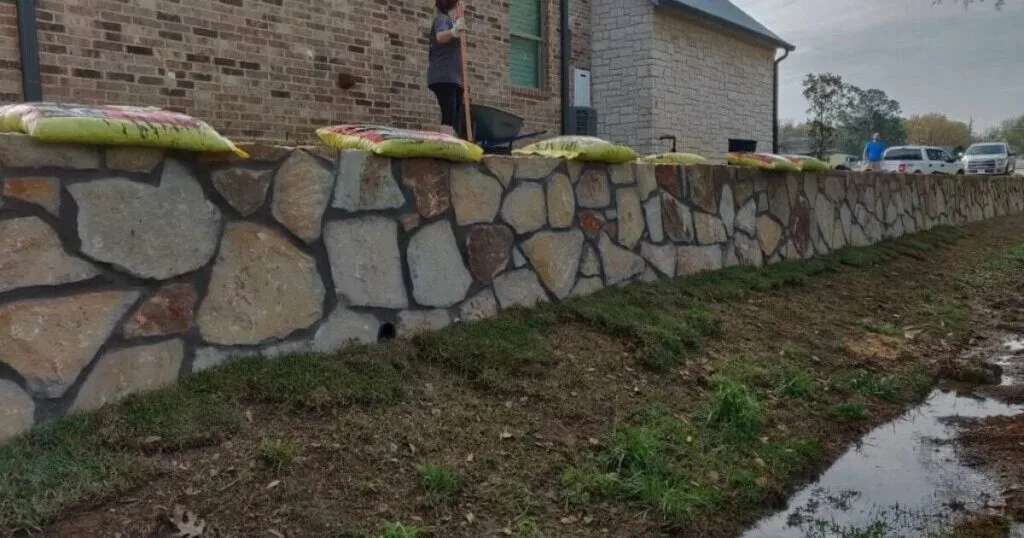 Newly built natural stone retaining wall along a brick home in Grapevine TX, with drainage weep hole and crew finishing backfill
