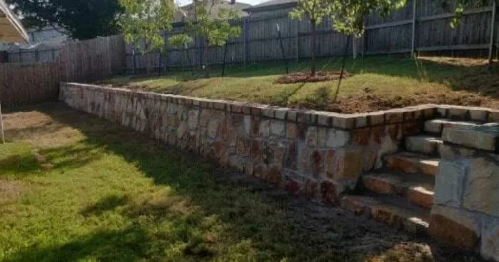 Natural stone retaining wall with built-in staircase and decorative cap separating tiered backyard levels in DFW residential property