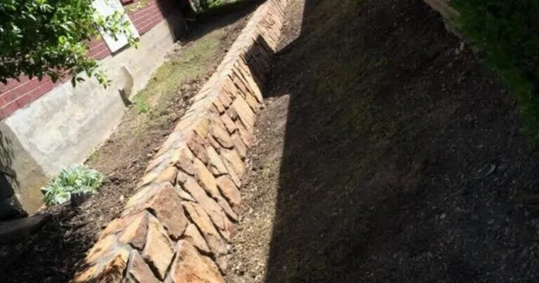 Newly built natural stone retaining wall along a residential yard slope beside a brick home, controlling erosion and grading in DFW clay soil.