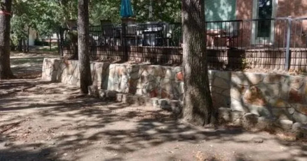 Natural stone retaining wall replacing old cross ties along a shaded residential yard in Highland Village TX, with mature trees and wrought-iron fence above