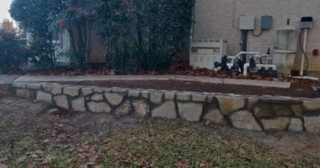 Curved natural stone retaining wall replacing a cross-tie flowerbed wall beside a brick home in Carrollton TX, with fresh soil bed and pool equipment visible.