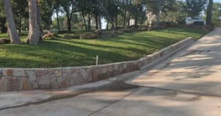 Natural stone retaining wall along a concrete driveway replacing old cross-tie wall, with graded lawn and mature trees, Dallas TX