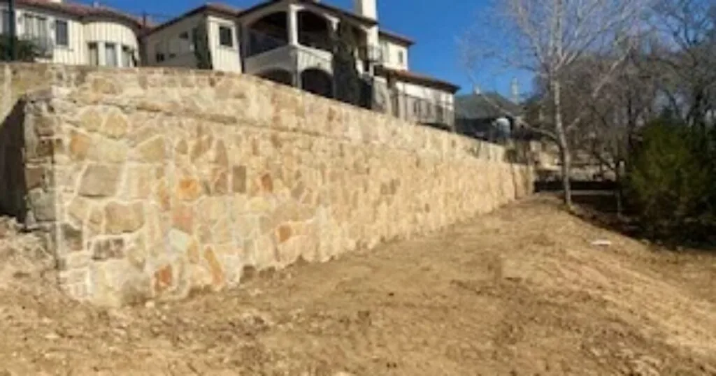 Large newly built natural stone retaining wall supporting a hillside residential property in DFW, with graded soil below