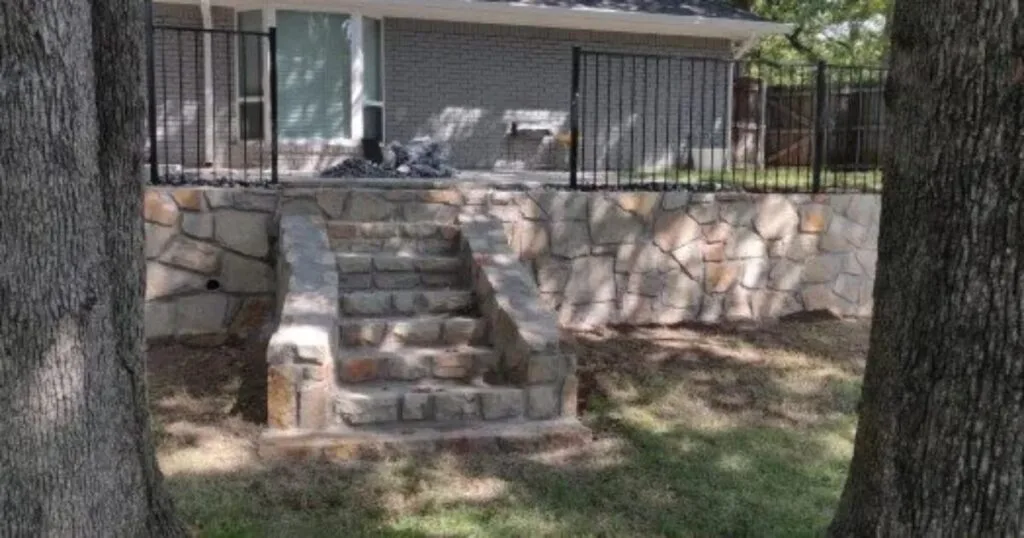 Natural stone retaining wall with built-in steps replacing old cross ties at a Carrollton TX home, topped with black iron railing