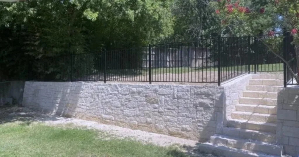 Completed natural stone retaining wall with built-in steps and black iron fence topper in a Dallas backyard