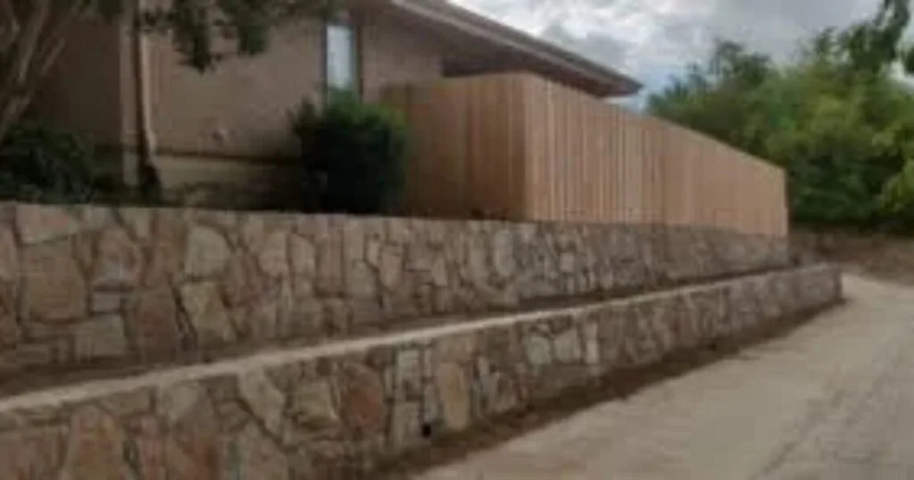 Two-tier natural stone retaining wall with cedar privacy fence atop, alongside residential driveway in Carrollton, TX