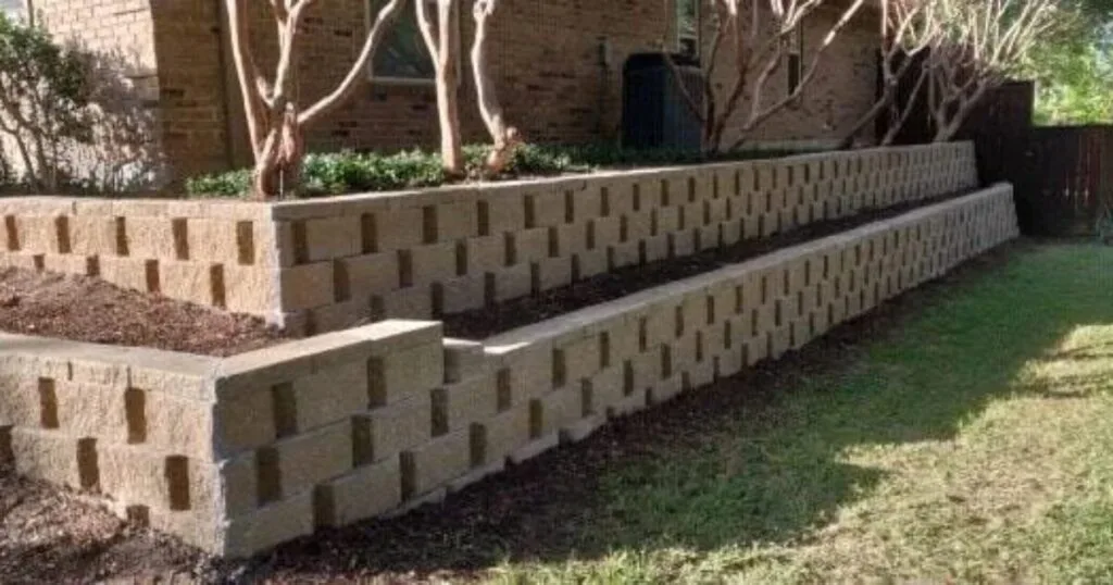 Two-tier concrete block retaining wall replacing old cross ties in a residential backyard in Arlington, TX, with planted beds and lawn